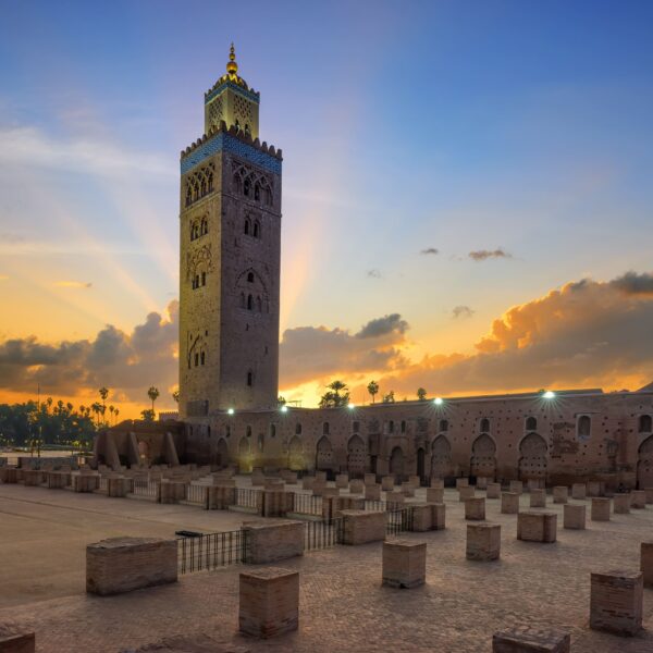 Uma vista serena do nascer do sol sobre a Mesquita Koutoubia em Marraquexe, Marrocos. O céu apresenta uma gradação de cores quentes à medida que o sol nasce, lançando um brilho suave sobre a estrutura histórica. Em primeiro plano, filas de pilares quadrados de pedra criam um padrão que conduz à mesquita. A imagem capta a essência da arquitetura marroquina e pode ser associada a debates sobre o mercado imobiliário marroquino e o custo de vida, realçando os elementos culturais e históricos que podem influenciar estes sectores.