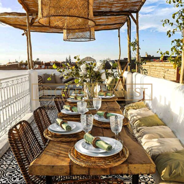 Charmante salle à manger du Riad sur un toit-terrasse avec un parasol en rotin, une table en bois pour le repas et une longue banquette avec des coussins.