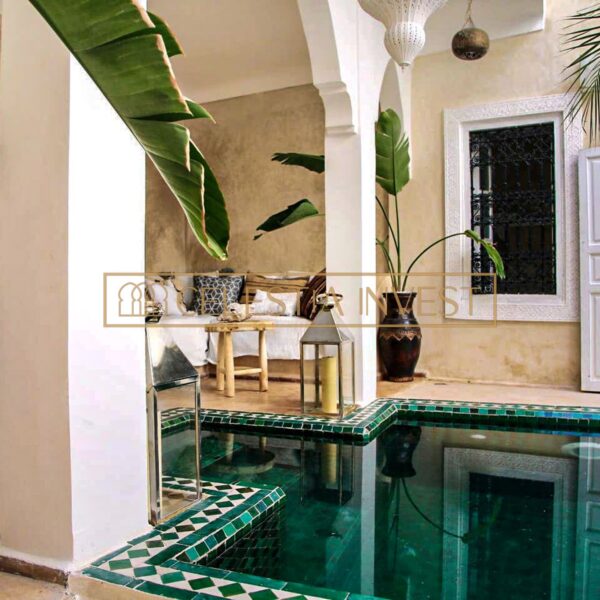 Un acogedor salón de Riad marroquí con vistas a la piscina del patio, adornado con exuberantes plantas verdes y una decoración elegante.