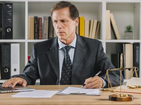 Un homme d'âge mûr travaillant sur un ordinateur portable tout en examinant un document dans un bureau de style juridique.
