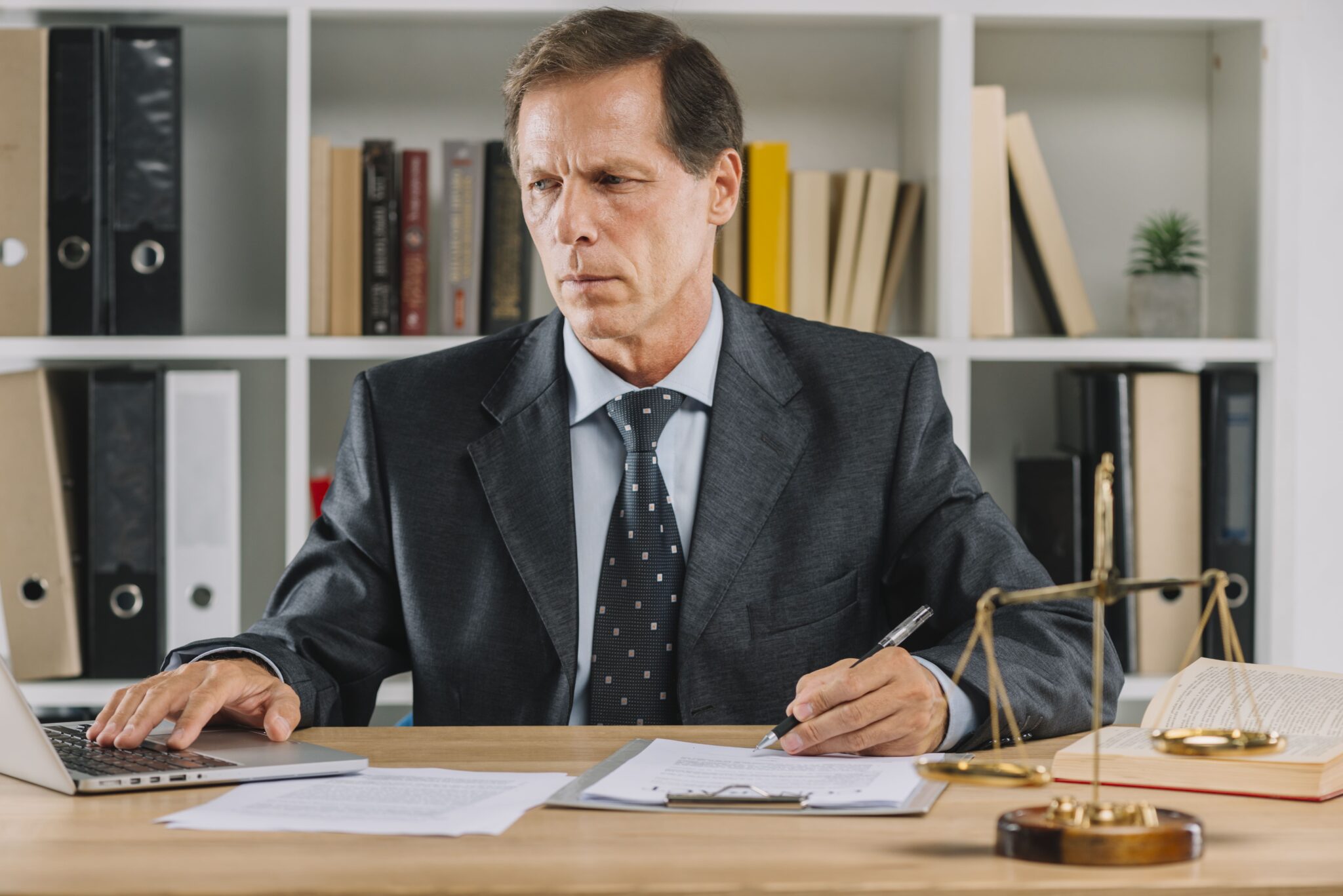 A middle-aged man working on a laptop while examining a document in a legal-style office.